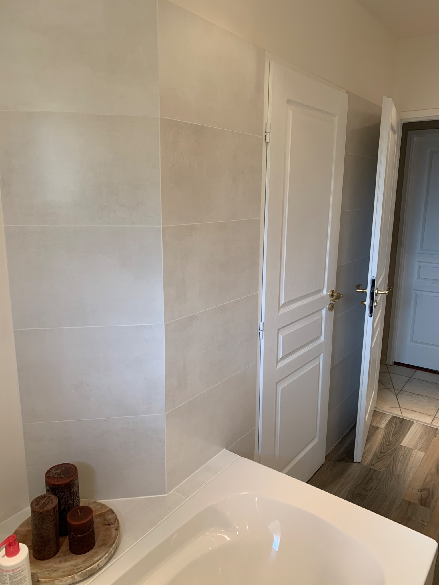 Jasne, poziome płytki na ścianie łazienki, biała wanna z widocznymi trzema brązowymi świecami na drewnianej podstawce, białe drzwi łazienkowe z widoczną podłogą z paneli w odcieniach brązu.