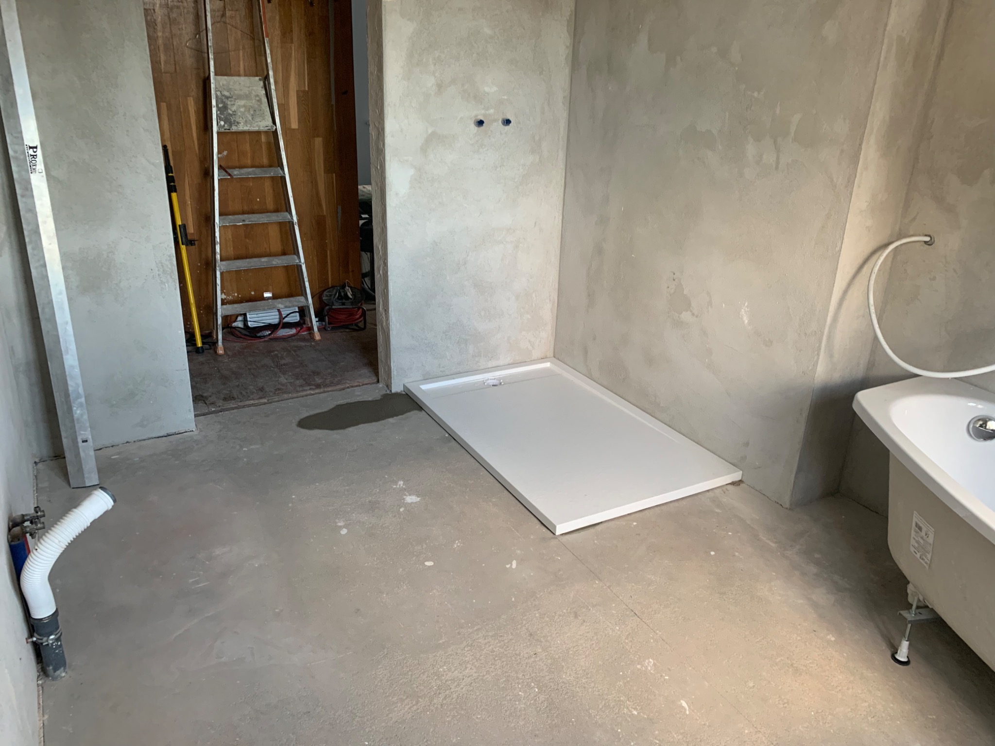 Łazienka w trakcie prac: surowe ściany, wylewka na podłodze, osadzony brodzik i wanna, drabina w tle.