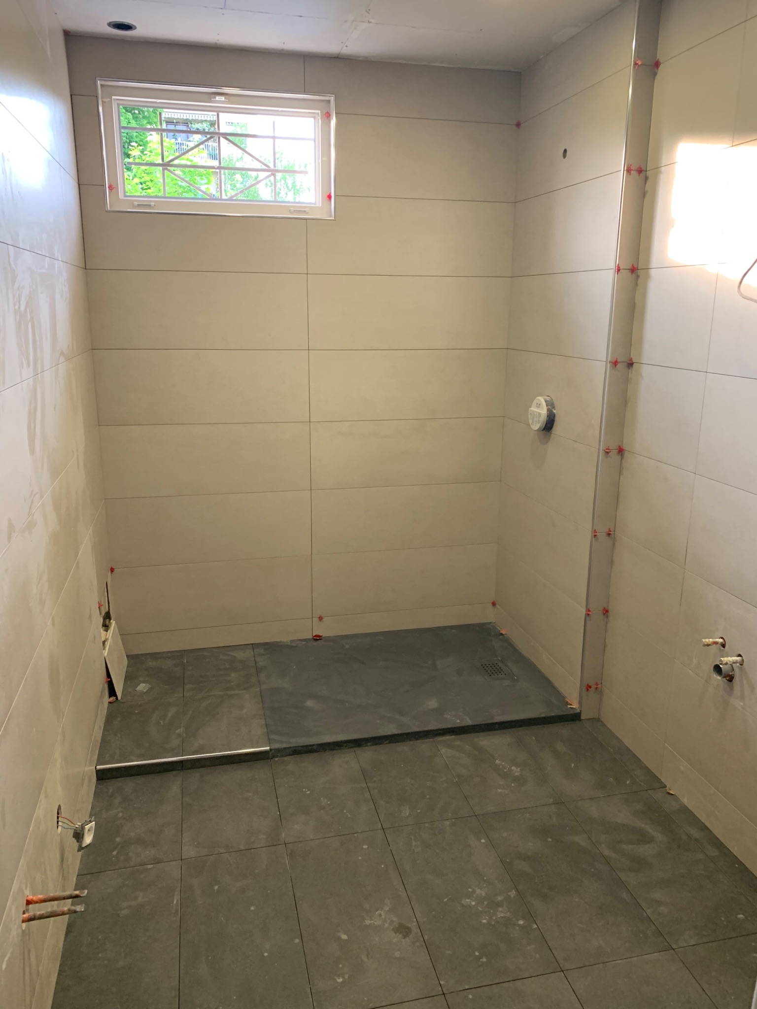 Wnętrze łazienki w trakcie układania beżowych płytek ściennych i grafitowych płytek podłogowych, widoczne krzyżyki dystansowe, okno z widokiem na zieleń.