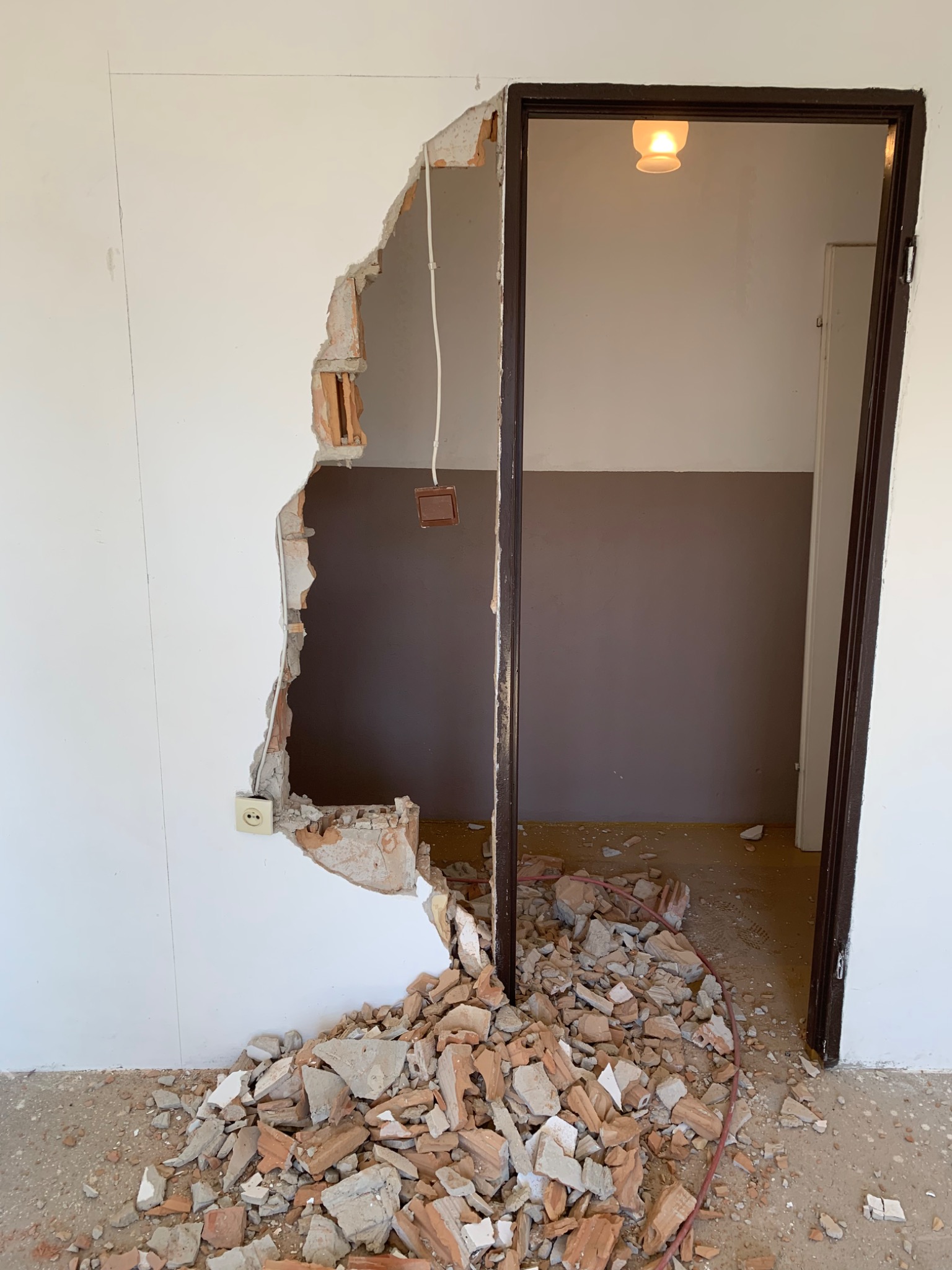 Uszkodzona ściana z widocznymi cegłami i instalacją elektryczną obok futryny drzwiowej, gruz na podłodze, widok na sąsiednie pomieszczenie z lampą sufitową.