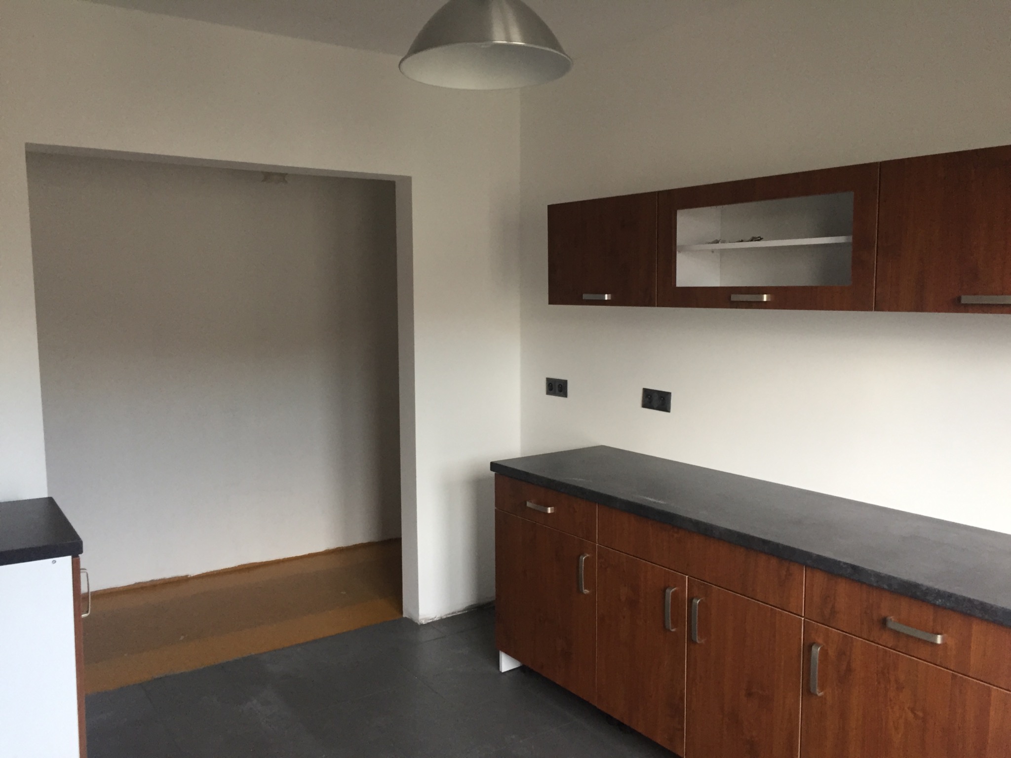 Kuchnia w trakcie prac wykończeniowych, widoczne brązowe szafki z szarym blatem, surowa podłoga i otwór w ścianie do innego pomieszczenia.