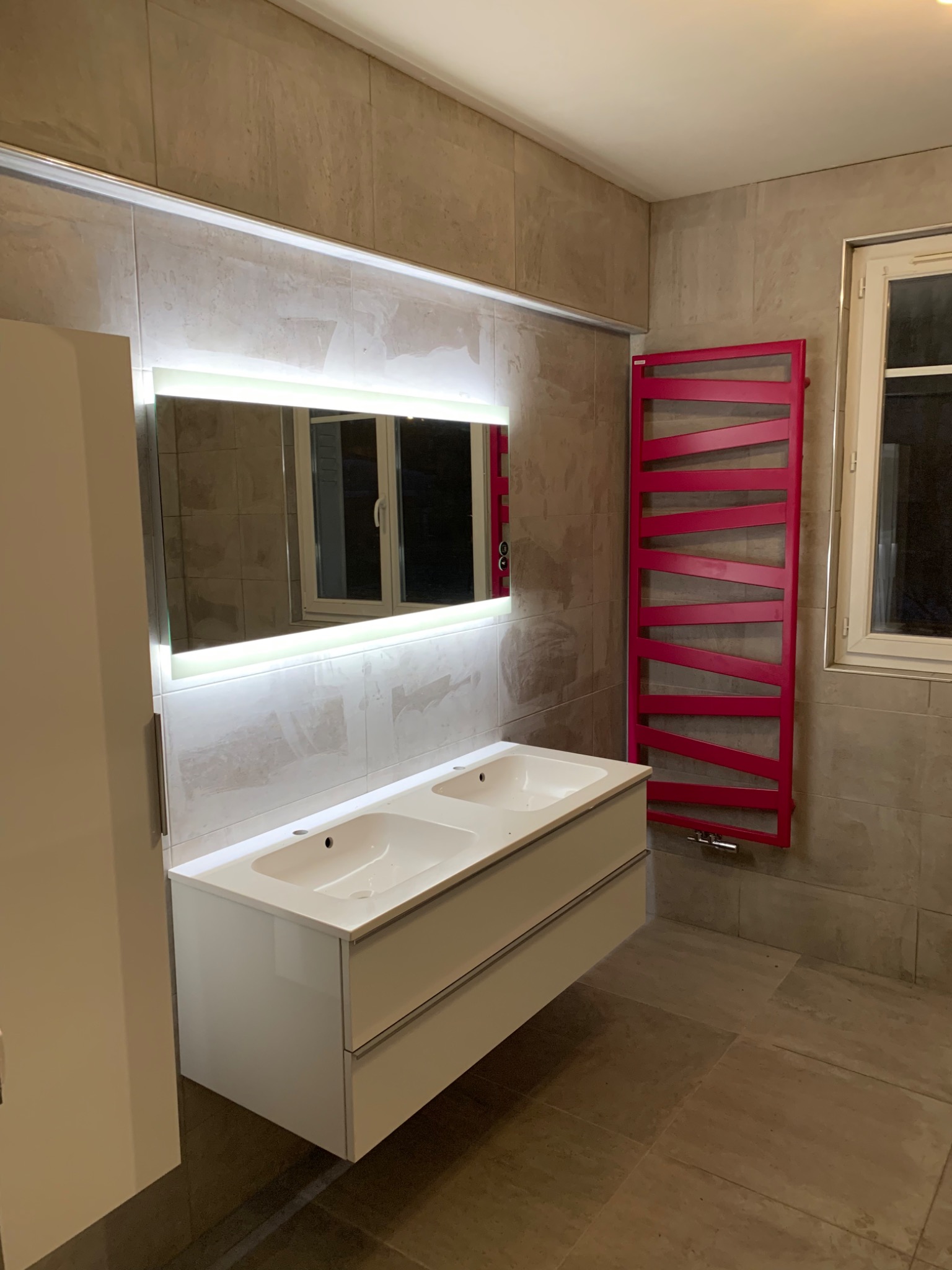 Nowoczesna łazienka z podwójną umywalką, lustrem z podświetleniem LED i fuksjowym grzejnikiem drabinkowym na tle beżowych płytek.