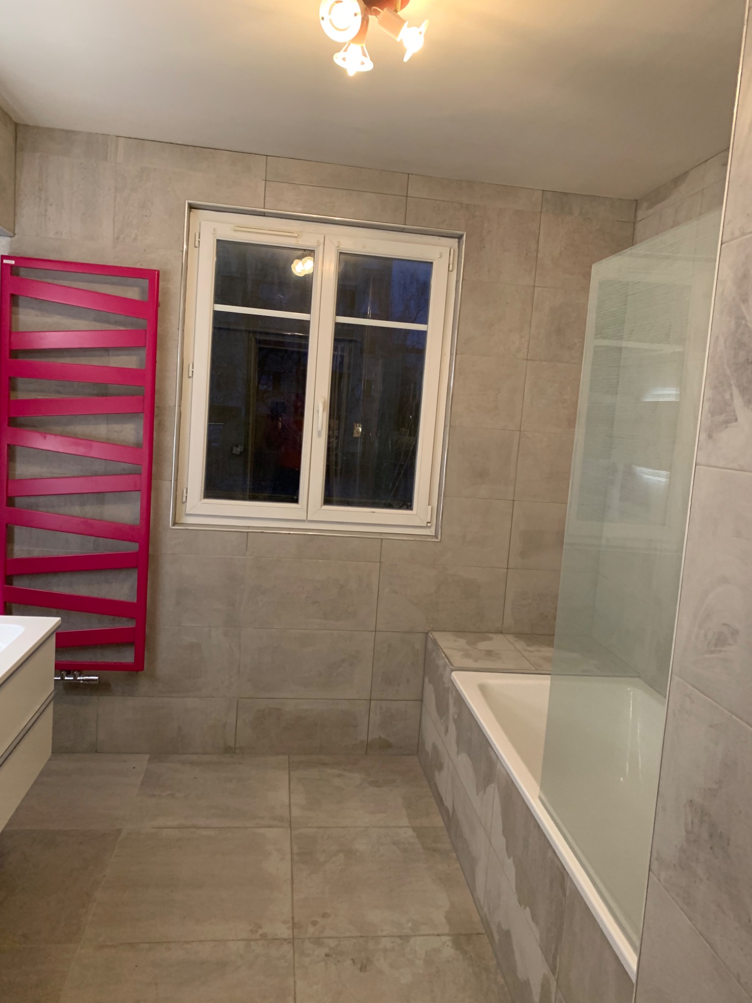 Wykończona łazienka z beżowymi płytkami, białą wanną z szklanym parawanem i fuksjowym grzejnikiem drabinkowym.