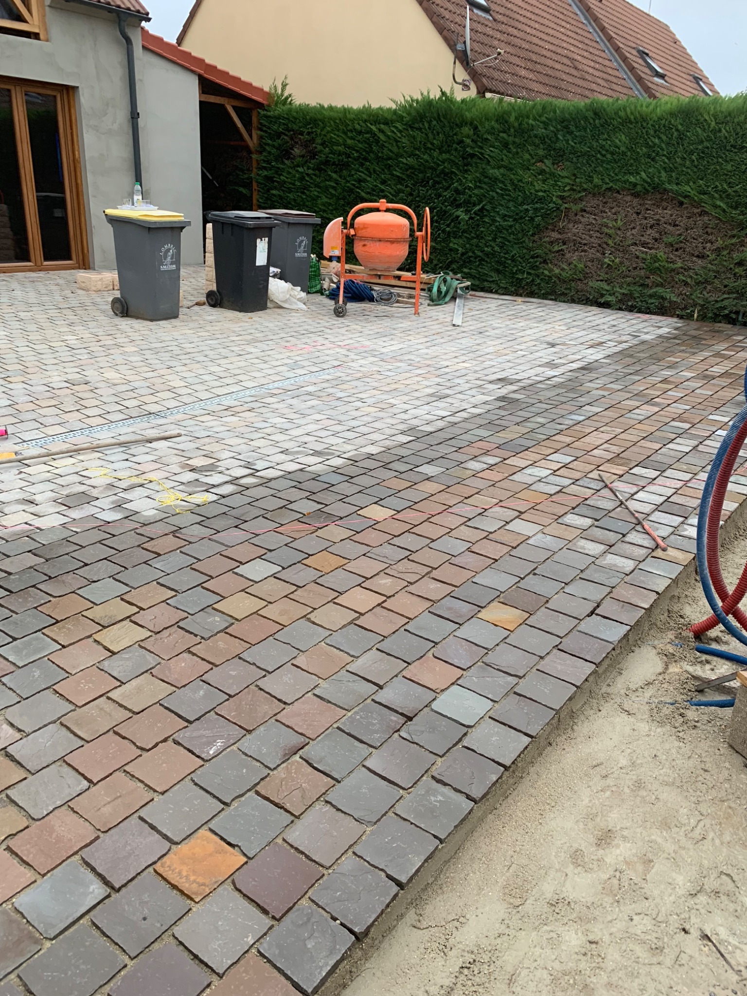 Układanie kostki brukowej zróżnicowanej kolorystycznie na podjeździe, widoczna betoniarka, ślady po wytyczaniu terenu, w tle dom jednorodzinny.