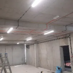 Surowy betonowy sufit z widocznymi przewodami elektrycznymi w kolorze pomarańczowym i czarnym, zamontowane lampy jarzeniowe, drabina aluminiowa i fragmenty sylwetek dwóch elektryków w kaskach...