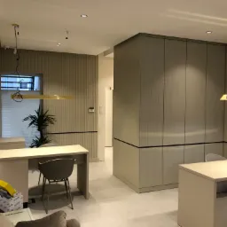 Nowoczesne wnętrze biurowe z beżowymi ścianami z pionowymi żłobieniami, minimalistycznymi biurkami i krzesłami w odcieniach szarości, oświetlone podłużnymi lampami sufitowymi, z widoczną rośliną...