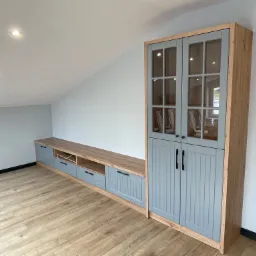 Zabudowa meblowa w pokoju na poddaszu: szafa z przeszklonymi drzwiczkami i szafka RTV z szufladami, w kolorze szaro-drewnianym, na drewnianej podłodze.