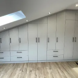 Biała szafa wnękowa z czarnymi uchwytami, dopasowana do skosu poddasza, oświetlenie punktowe w suficie, widoczne okno dachowe i fragment odkurzacza.