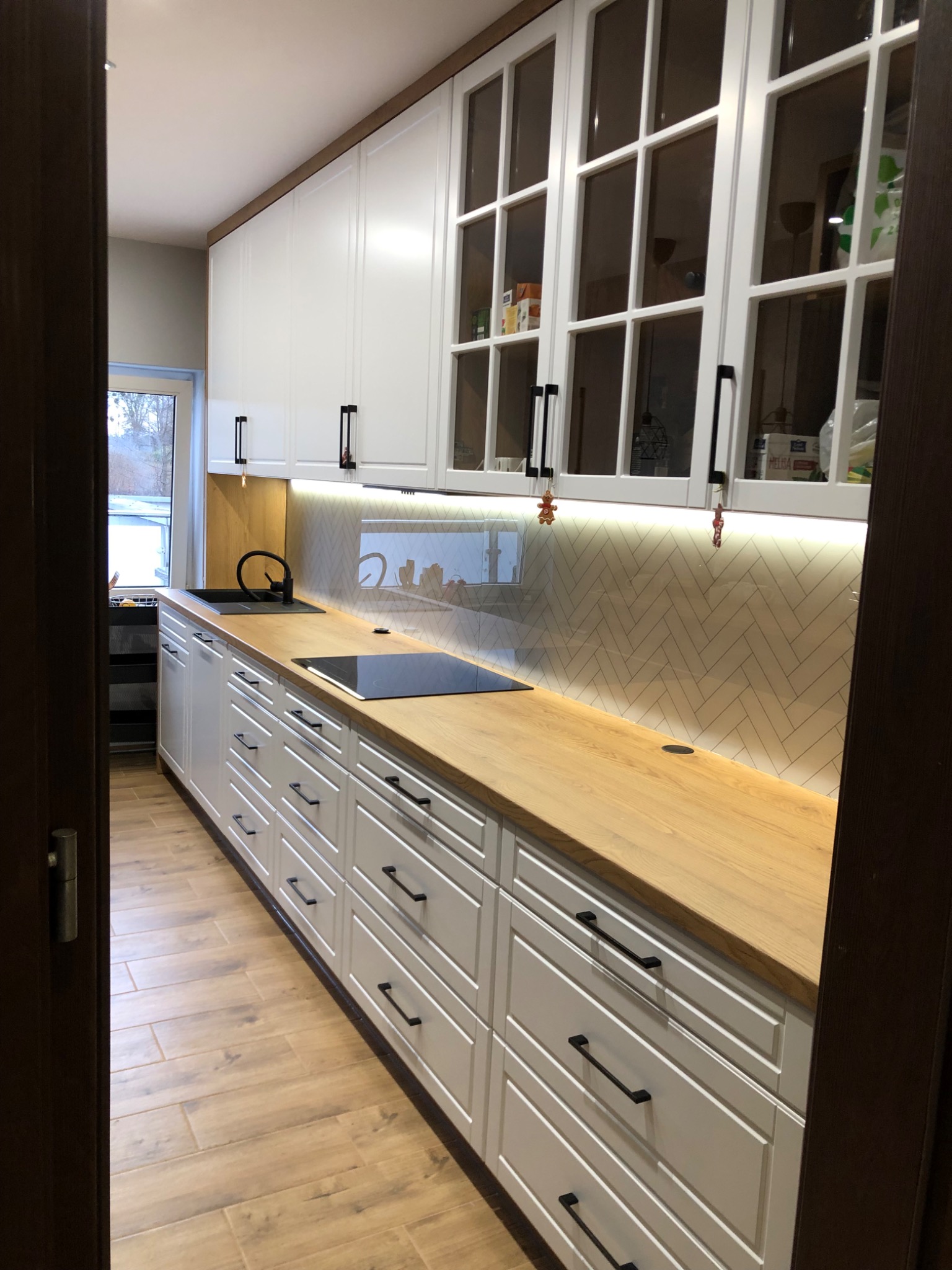 Nowoczesna kuchnia z białymi frontami, drewnianym blatem i podświetlanym backsplash'em w jodełkę, widziana z perspektywy wejścia.