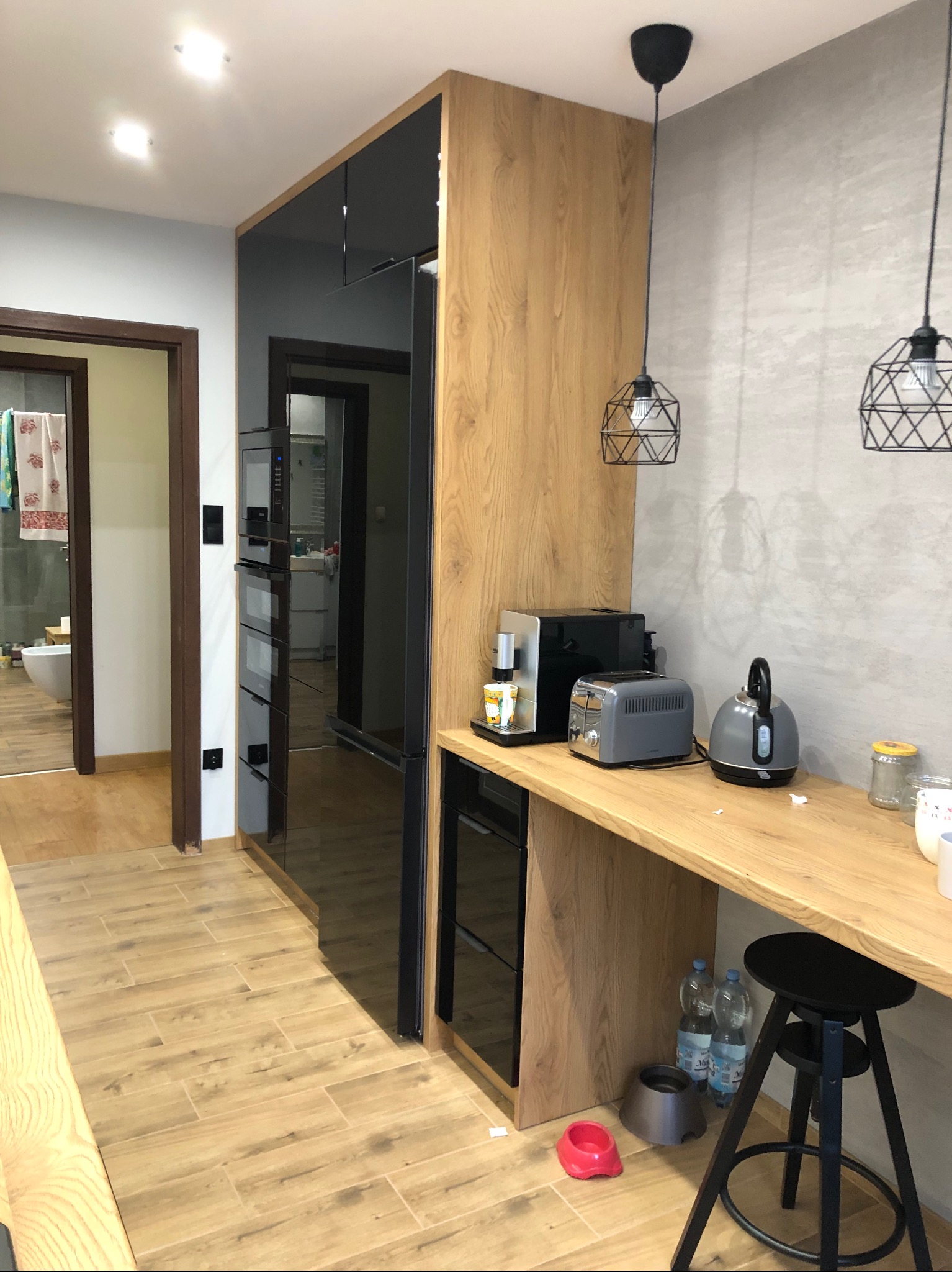 Nowoczesna kuchnia z drewnianymi frontami szafek, czarnymi elementami i blatem roboczym, oświetlona wiszącymi lampami z geometrycznymi kloszami. Widoczny sprzęt AGD, wysoki stołek barowy i podłoga...