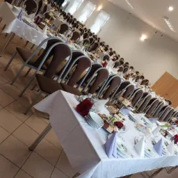 Sala bankietowa przygotowana do przyjęcia, długie stoły nakryte białymi obrusami, zastawa, dekoracje, krzesła z brązowymi oparciami ustawione w rzędach.