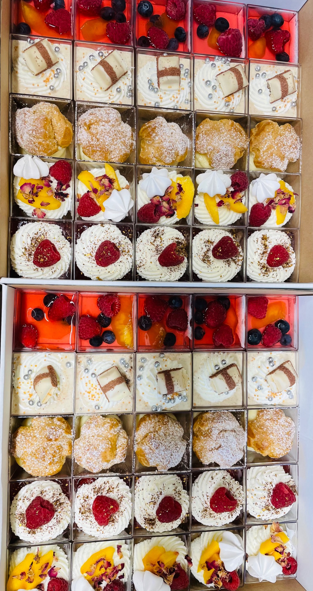 Zestaw różnorodnych deserów w przezroczystych pojemnikach, ułożonych w kartonowym pudełku, zawierający kremowe babeczki z owocami, galaretki z jagodami i malinami, oraz ptysie posypane cukrem pudrem.