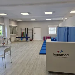 Jasne, przestronne wnętrze gabinetu rehabilitacyjnego z recepcją Bonumed, sprzętem do ćwiczeń, drabinkami, kozetką i zasłoniętym parawanem.