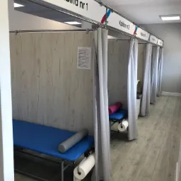 Wnętrze gabinetu z kilkoma stanowiskami do masażu lub rehabilitacji, każde oddzielone zasłonami i oznaczone numerem, wyposażone w niebieskie leżanki i rolki do ćwiczeń.