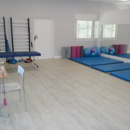NZOZ Centrum Rehabilitacji BonuMed - Jasne wnętrze gabinetu z drabinkami gimnastycznymi, niebieskim kozetką, stolikiem z przyrządami i niebieskimi matami pod ścianą z lustrem.