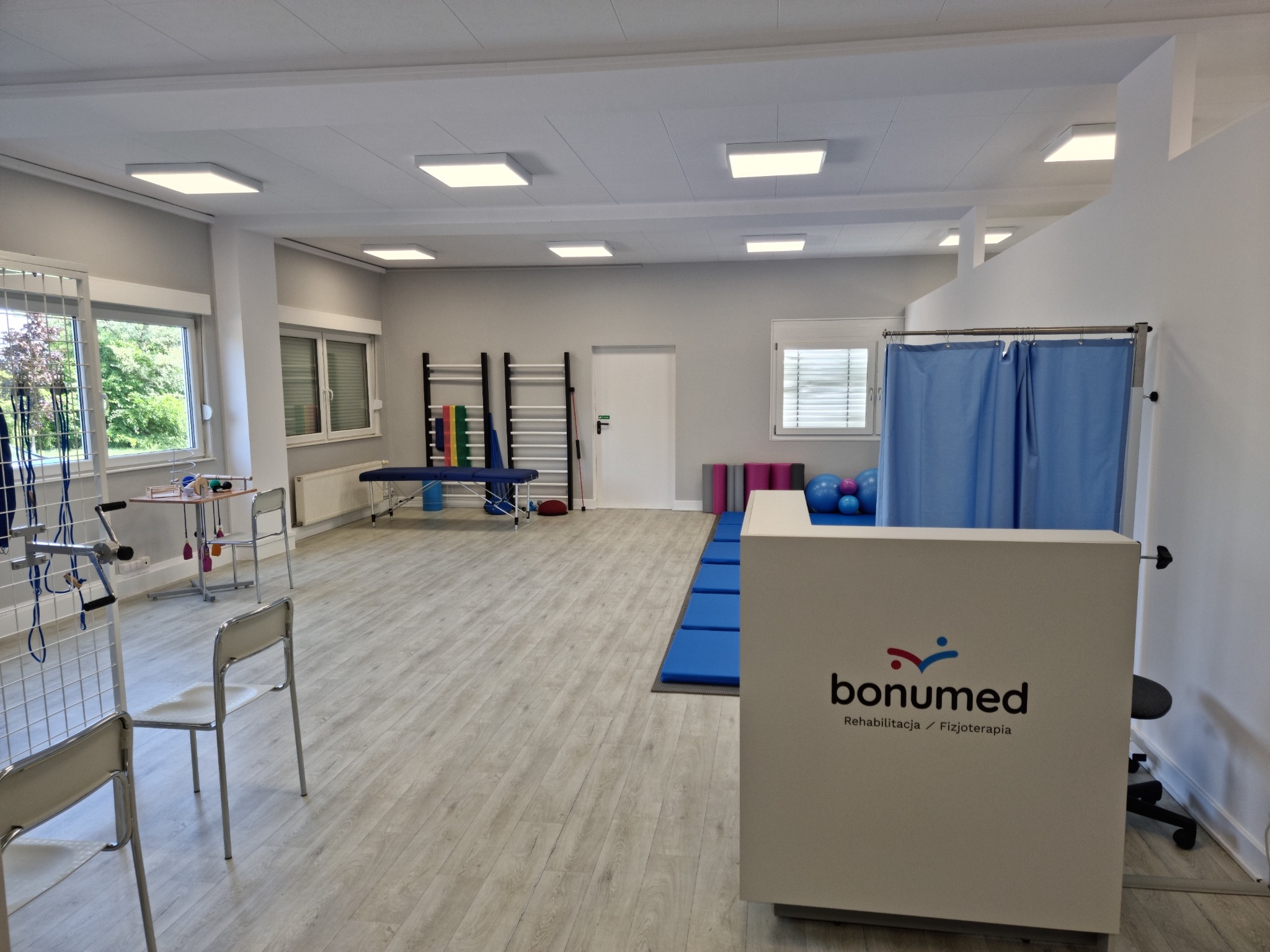 Jasne, przestronne wnętrze gabinetu rehabilitacyjnego z recepcją Bonumed, sprzętem do ćwiczeń, drabinkami, kozetką i zasłoniętym parawanem.