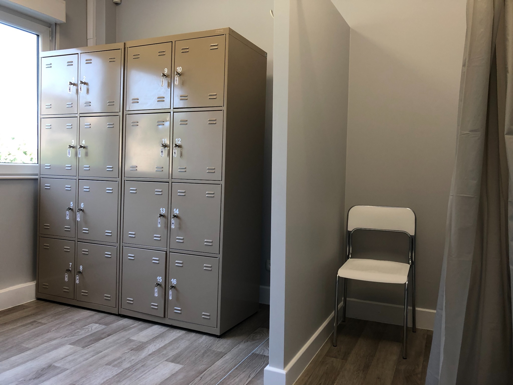 Szereg szafek depozytowych z numerami, ustawionych w jasnym pomieszczeniu z drewnopodobną podłogą, obok białe krzesło i zasłona.