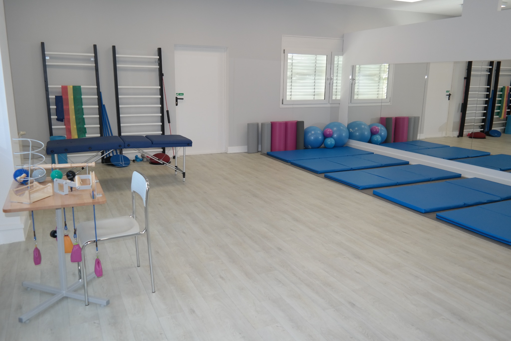Jasne wnętrze gabinetu z drabinkami gimnastycznymi, niebieskim kozetką, stolikiem z przyrządami i niebieskimi matami pod ścianą z lustrem.