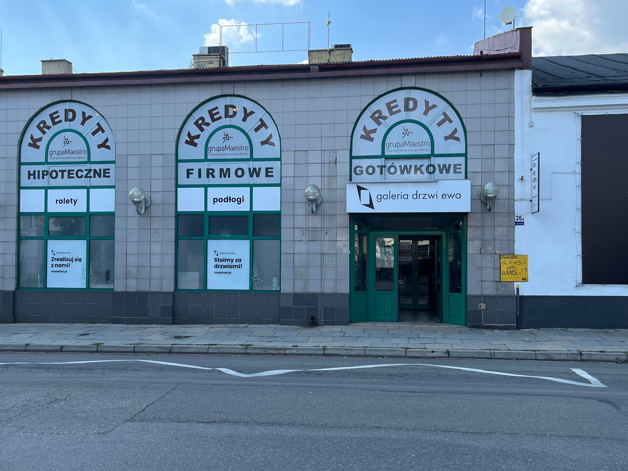 Fasada budynku z trzema wejściami oznaczonymi jako Kredyty Hipoteczne, Firmowe i Gotówkowe Grupy Maestro, z dodatkową informacją o sprzedaży rolet, podłóg i Galerii Drzwi Ewa.