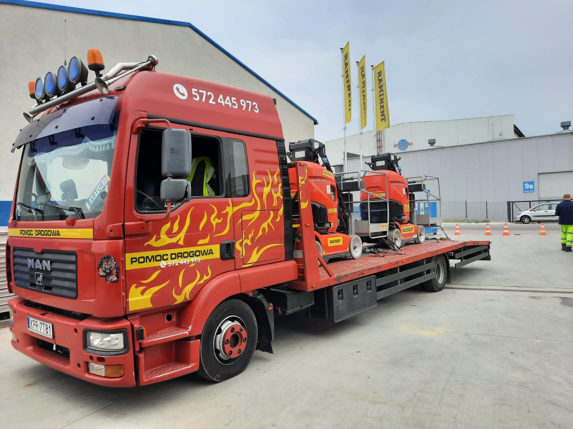 Czerwona laweta MAN z żółtymi płomieniami i napisem 'POMOC DROGOWA', przewożąca dwie pomarańczowe podnośniki koszowe firmy Ramirent na platformie, zaparkowana na betonowym placu przed halą...