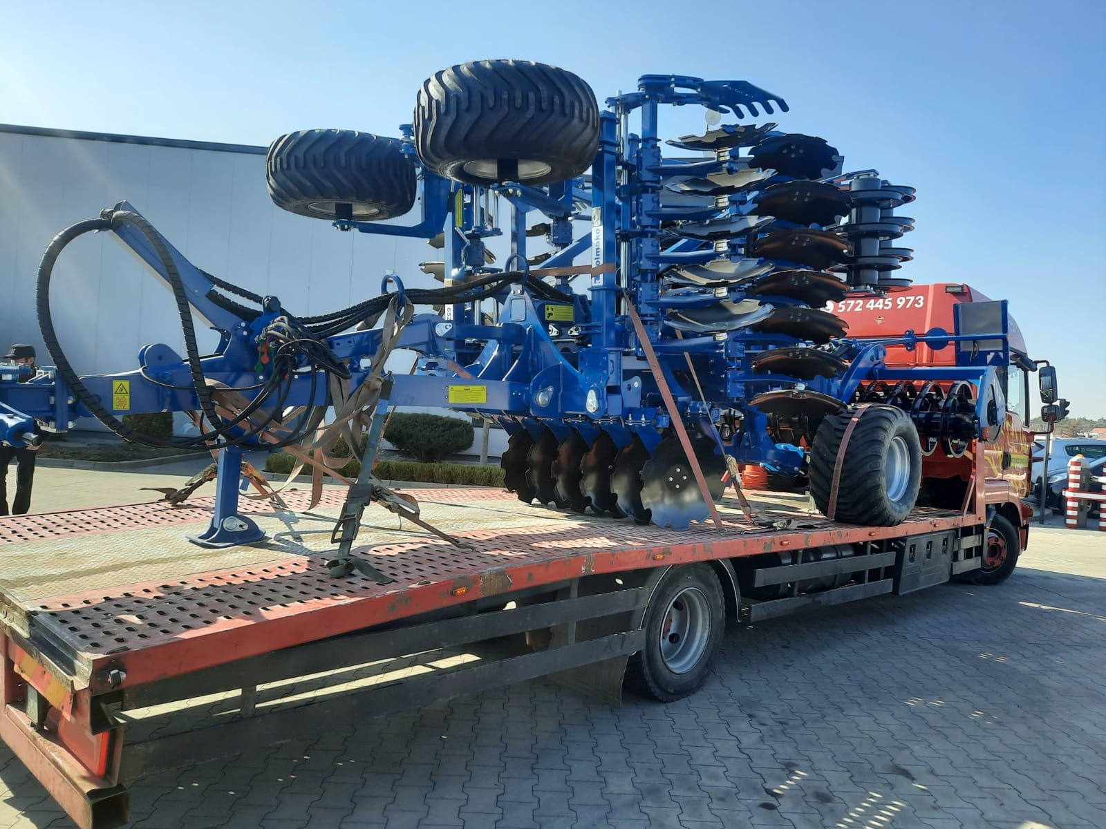 Niebieski agregat uprawowy marki Kverneland na czerwonej platformie niskopodwoziowej ciężarówki, zabezpieczony pasami transportowymi, na tle budynku przemysłowego i błękitnego nieba.