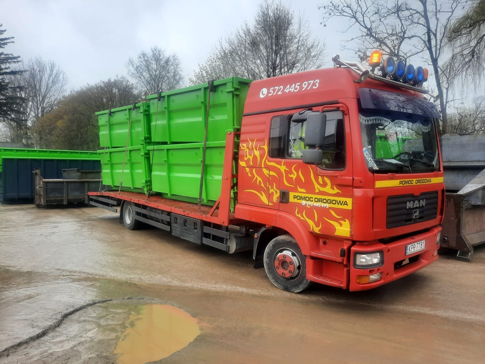 Czerwona ciężarówka MAN z platformą przewożąca dwa duże, zielone kontenery na tle drzew i kałuż na placu. Na kabinie widoczne logo 'Pomoc Drogowa' i numer telefonu.