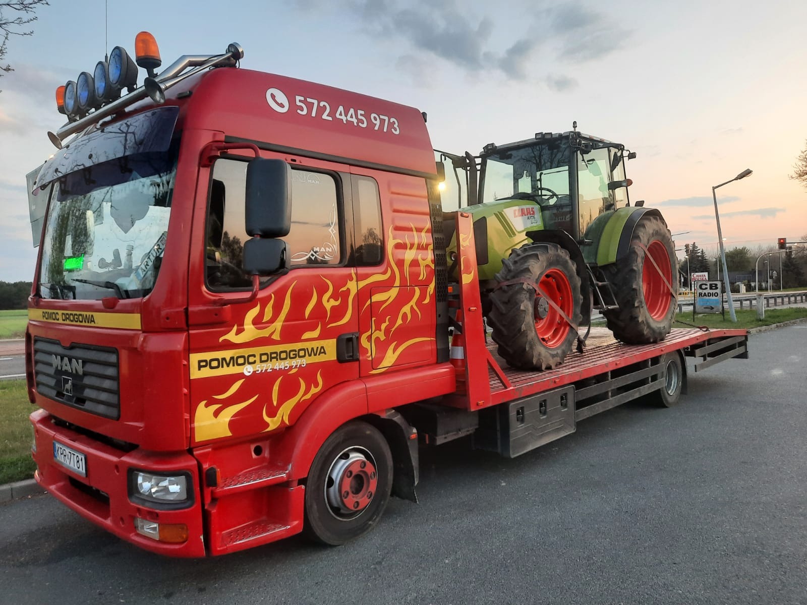 Czerwona laweta z grafiką płomieni transportuje zielony traktor Claas Axos na platformie. Na lawecie widoczne numery telefonu i napis 'Pomoc Drogowa'.