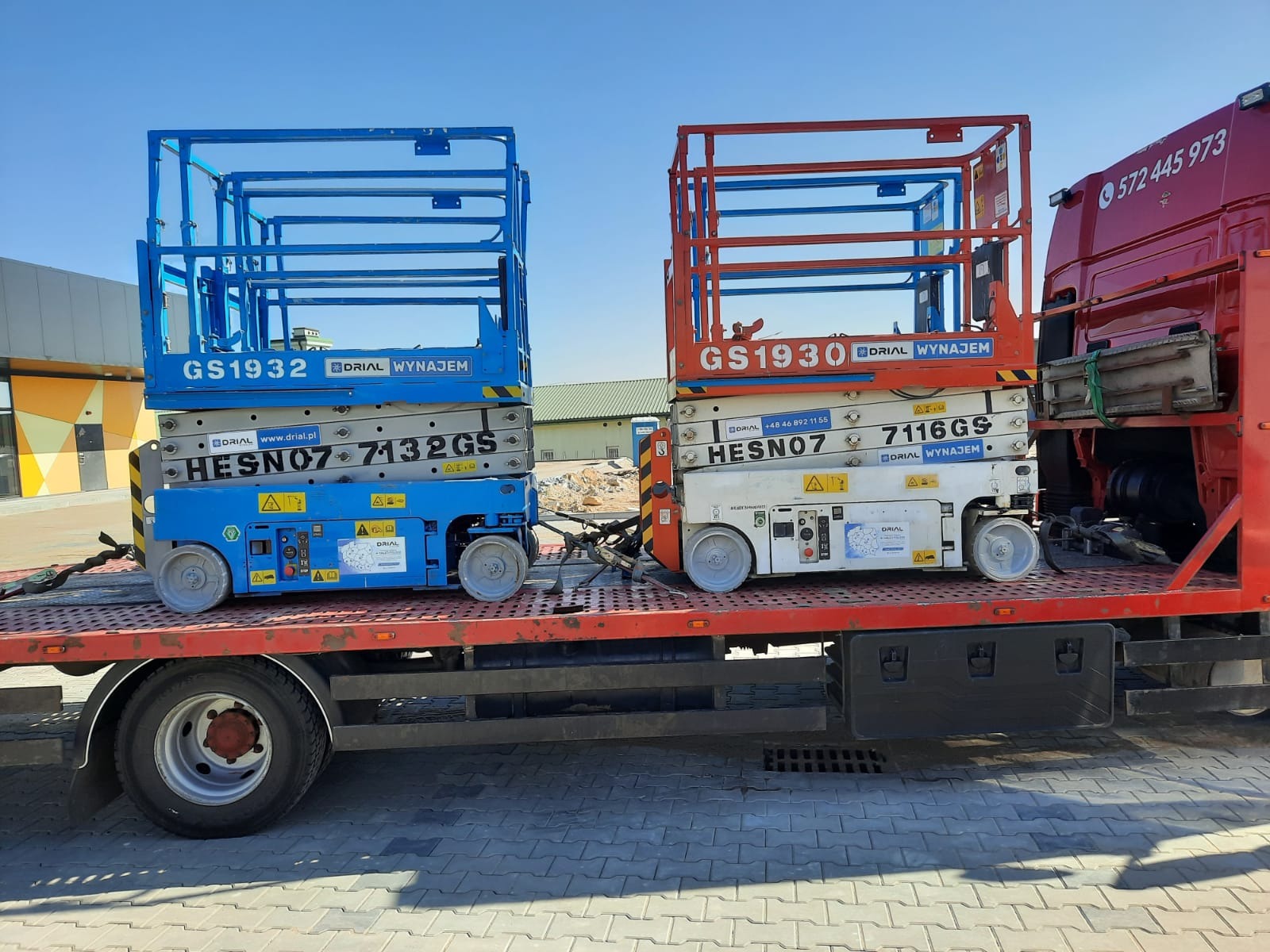 Dwa podnośniki nożycowe, niebieski GS1932 i pomarańczowy GS1930, zabezpieczone na platformie czerwonej ciężarówki, gotowe do przewozu. Widoczny fragment kabiny ciężarówki z numerem telefonu.
