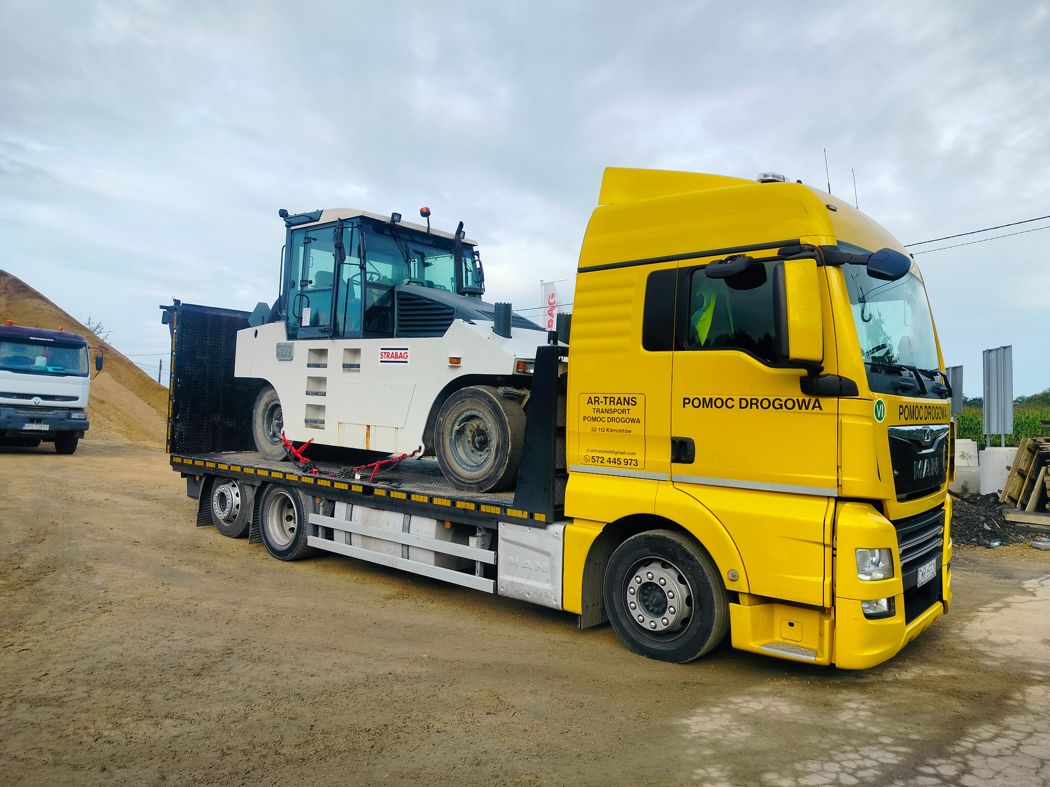 Żółta laweta z napisem 'POMOC DROGOWA' przewozi biały, specjalistyczny pojazd STRABAG zabezpieczony czerwonymi pasami transportowymi.