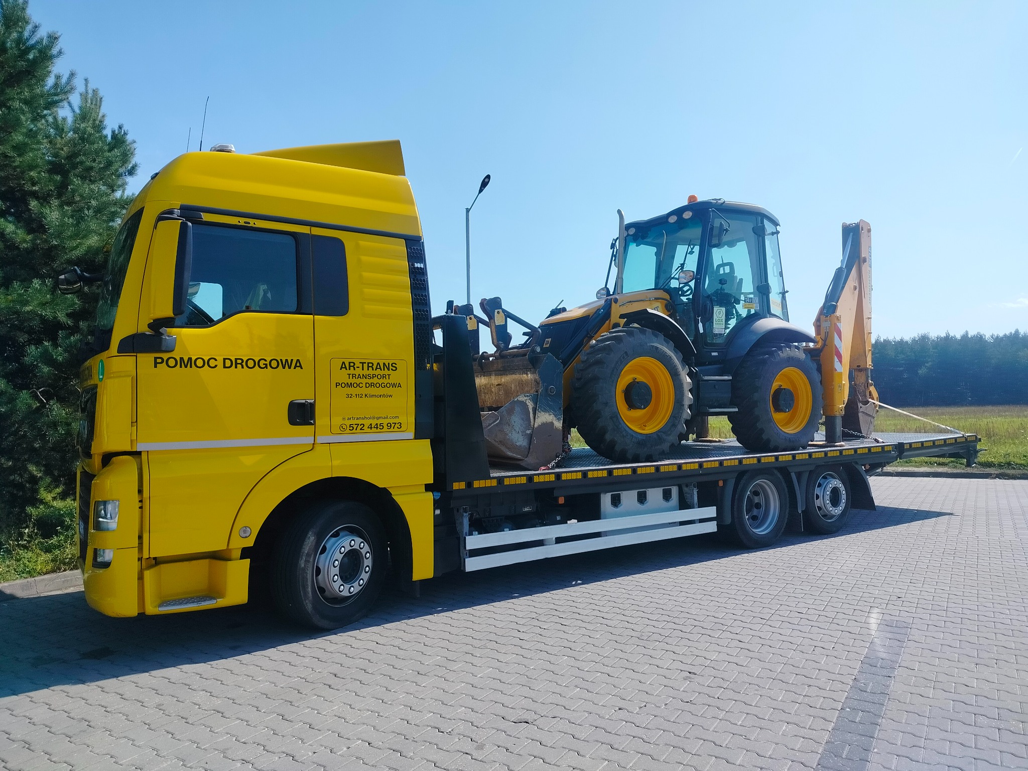 Żółta laweta z napisem 'Pomoc Drogowa' przewozi żółto-czarną koparko-ładowarkę na tle błękitnego nieba i zieleni.