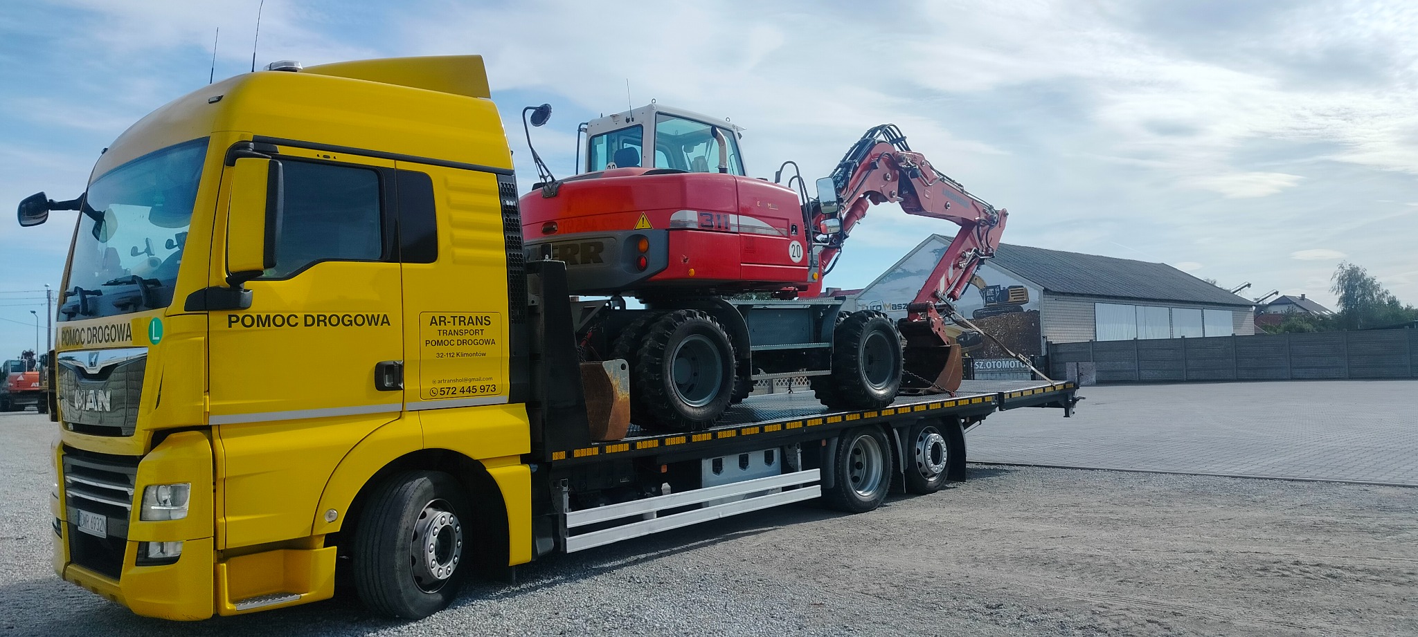 Żółta laweta z napisem 'Pomoc Drogowa' przewozi czerwony, kołowy bager marki Liebherr na platformie. Widoczne logo firmy AR-TRANS na kabinie. Tło stanowi utwardzony plac i budynek z muralem...