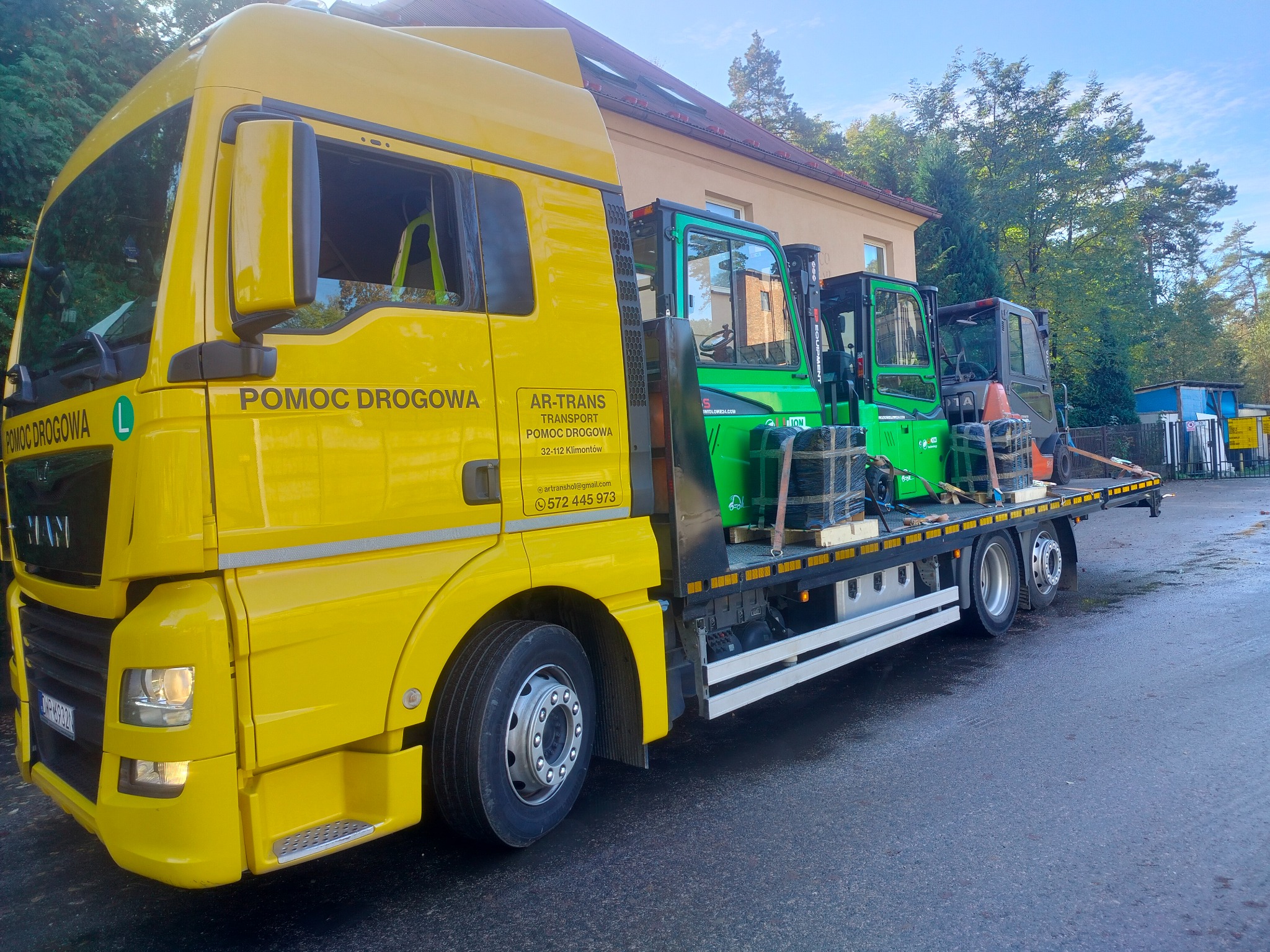 Żółta laweta pomocy drogowej AR-Trans przewozi dwa zielone wózki widłowe na platformie, widok z boku na tle budynku i drzew.