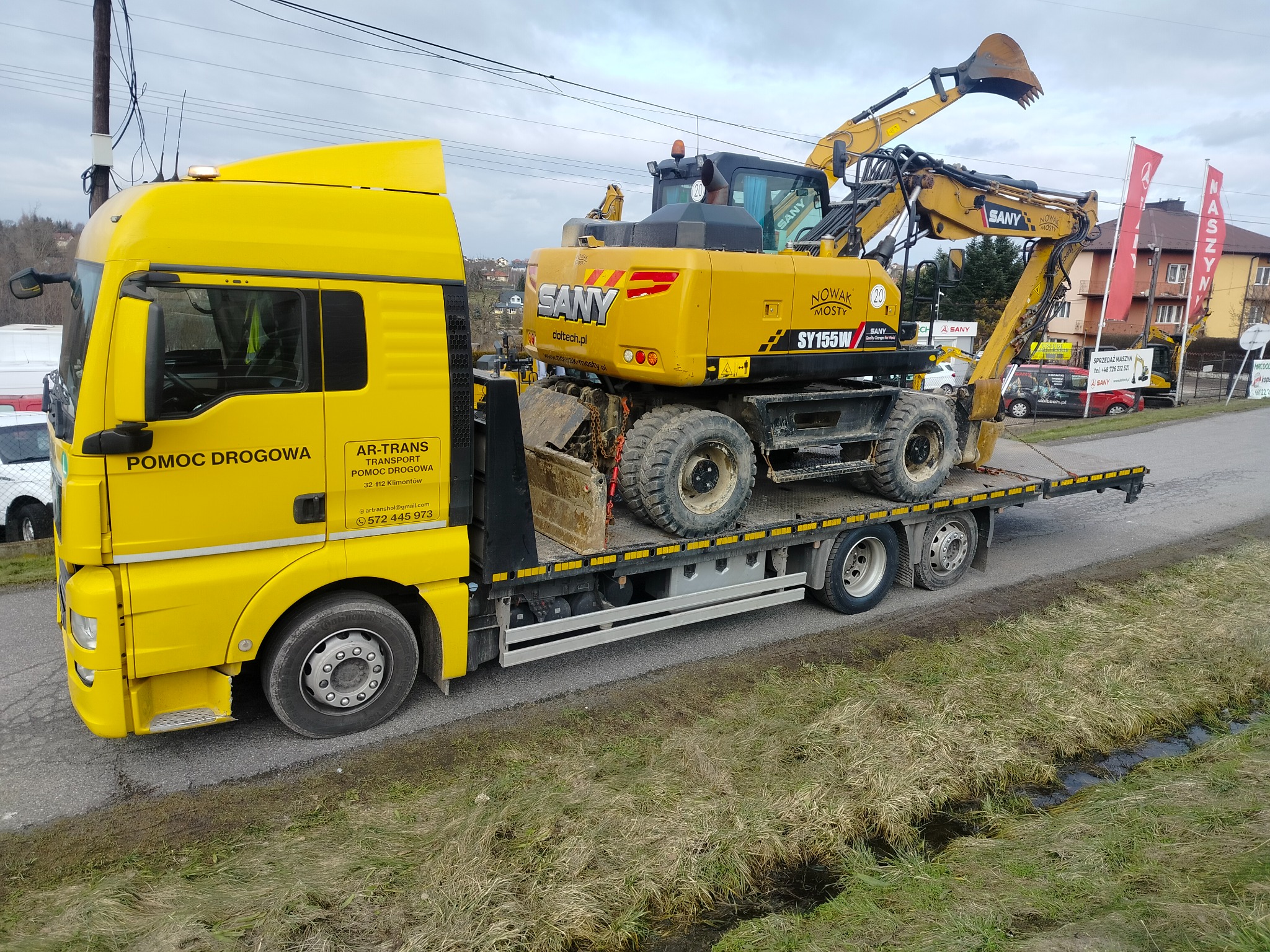 Żółta laweta z dźwigiem SANY SY155W na platformie, widok z boku na tle zabudowań i masztów reklamowych.