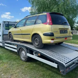 Żółty samochód kombi marki Peugeot przewożony na lawecie, widok z tyłu, zielona trawa w tle.