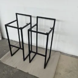 Dwie surowe, czarne, metalowe ramy krzeseł barowych, bez siedzisk, ustawione na tekturze falistej w surowym wnętrzu z białą ścianą.