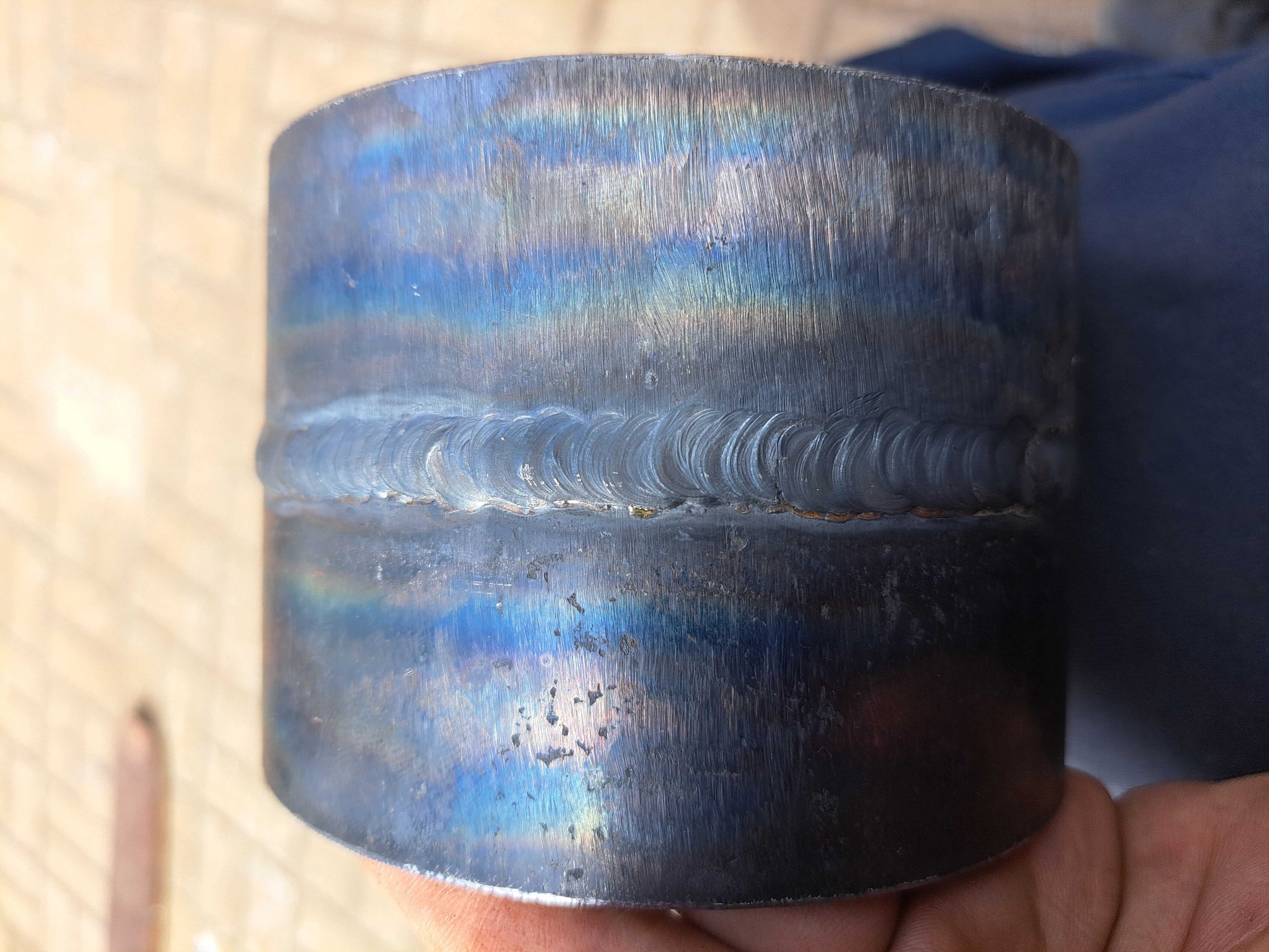 Metalowy cylinder z widocznym spoiną, z opalizującym zabarwieniem powierzchni w odcieniach niebieskiego i fioletu, trzymany w dłoni.