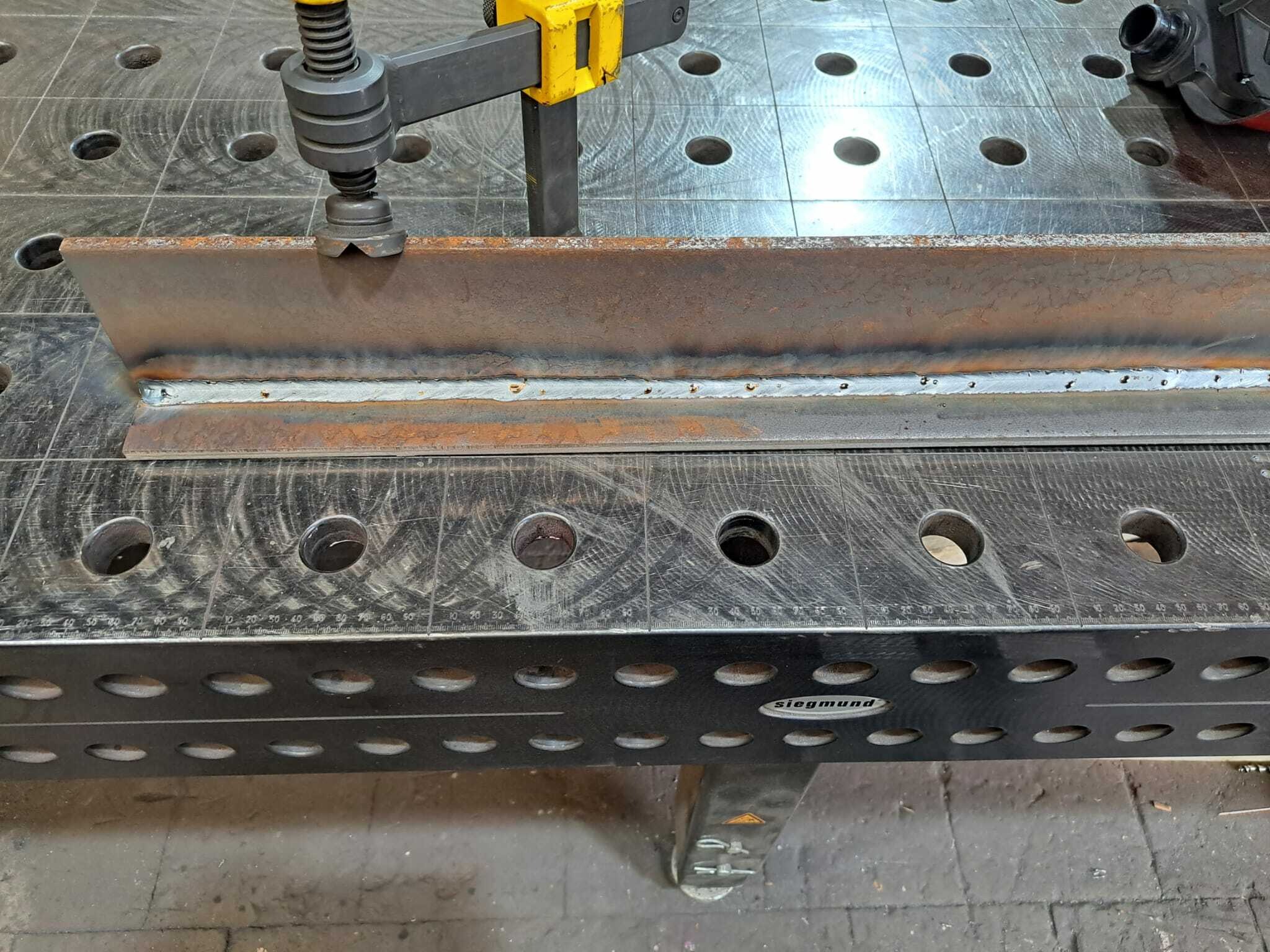 Spoina łącząca dwa elementy stalowe, dociskane żółtym ściskiem na perforowanym stole spawalniczym Siegmund.