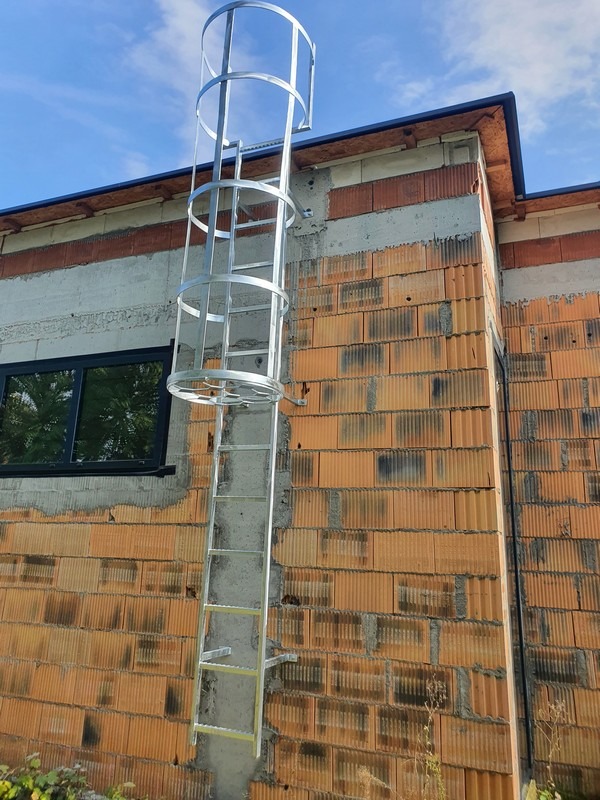 Zewnętrzna, metalowa drabina ewakuacyjna z koszem ochronnym, zamontowana na ścianie budynku z czerwonej cegły, widok z dołu, błękitne niebo w tle.