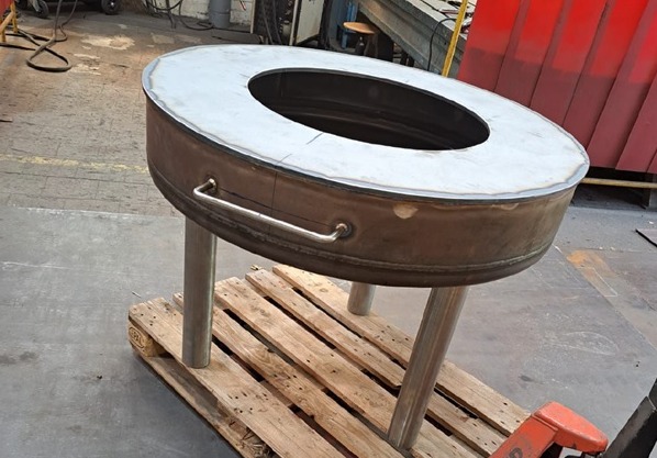 Owalny, spawany metalowy grill z uchwytem i trzema nogami, umieszczony na drewnianej palecie w hali produkcyjnej.