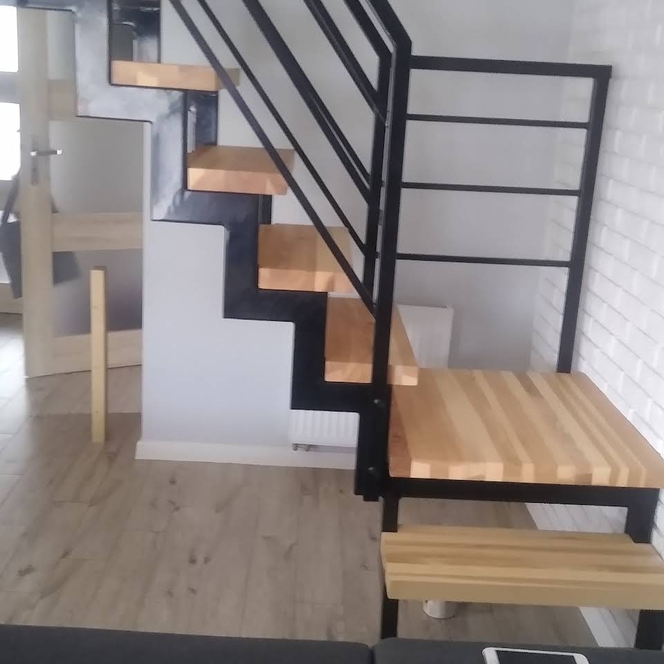 Schody stalowe z drewnianymi stopniami i czarną balustradą, zamontowane w nowoczesnym wnętrzu z białą cegłą na ścianie.