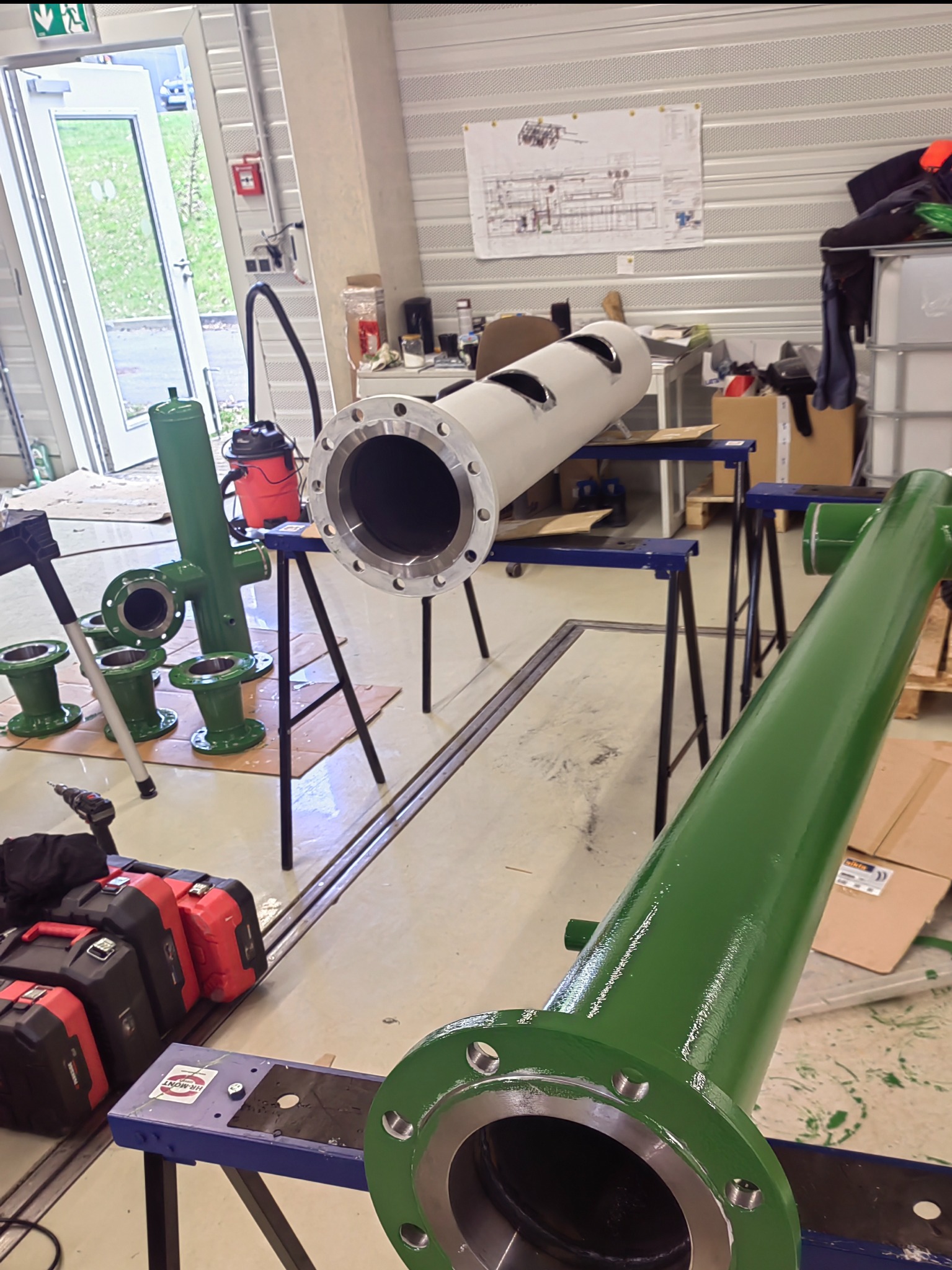 Elementy konstrukcji stalowej: rury z kołnierzami, pomalowane na zielono i szaro, leżące na kozłach w warsztacie, widoczne narzędzia i schemat na ścianie.