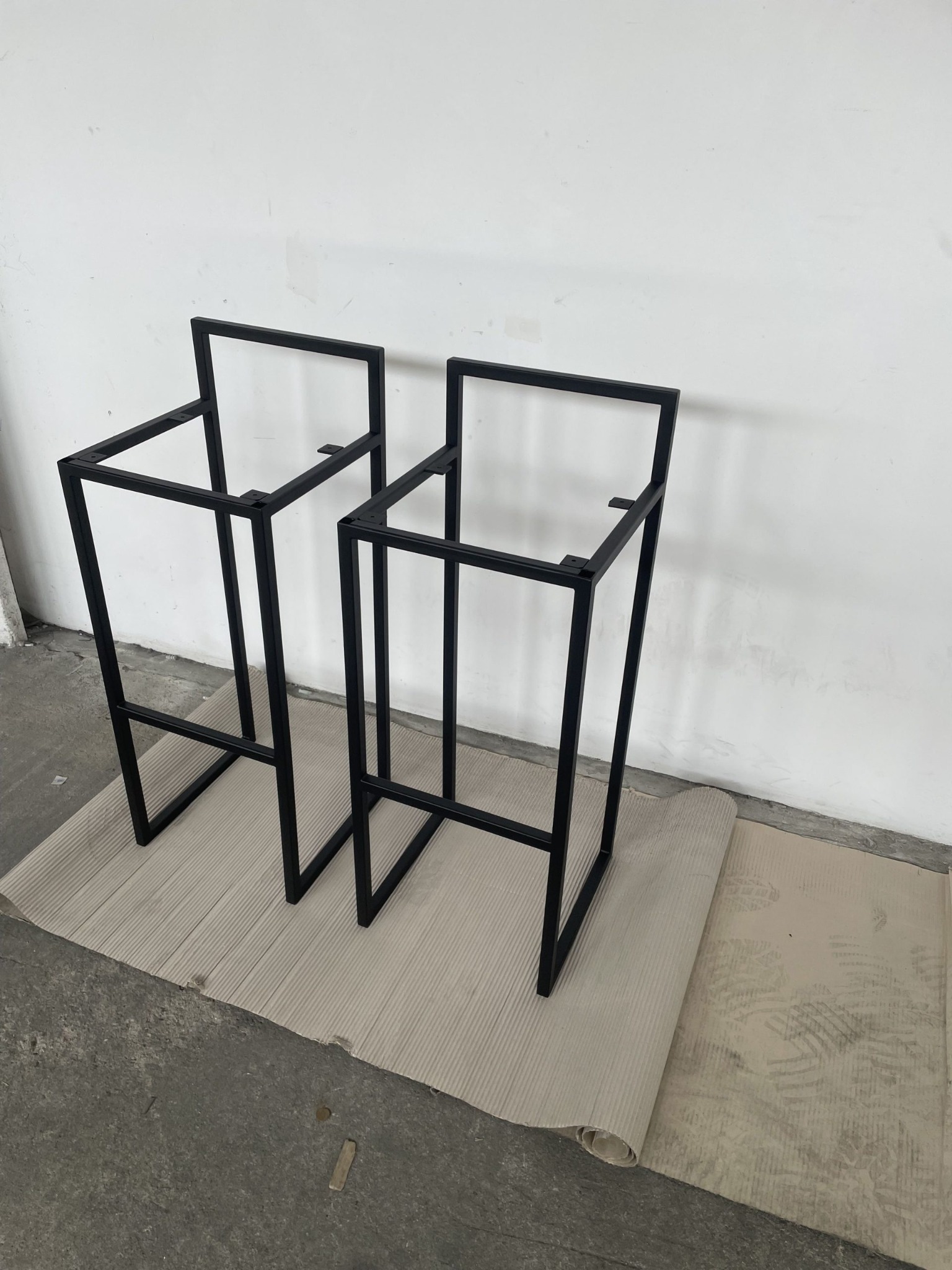 Dwie surowe, czarne, metalowe ramy krzeseł barowych, bez siedzisk, ustawione na tekturze falistej w surowym wnętrzu z białą ścianą.