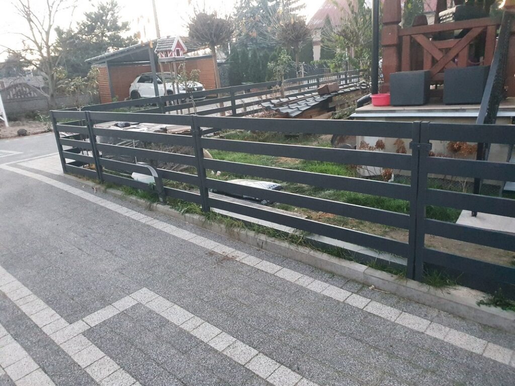 Nowoczesne, metalowe ogrodzenie posesji w kolorze grafitowym, z poziomymi szczeblami, widok z zewnątrz na kostkę brukową i fragment domu z drewnianym tarasem w tle.
