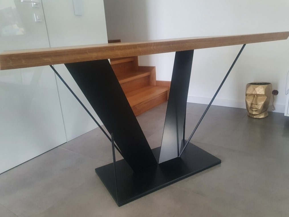 Stół konsola z blatem z jasnego drewna i czarną, geometryczną podstawą ze stali, umieszczony w nowoczesnym wnętrzu z widocznymi schodami i złotą dekoracją w kształcie głowy.