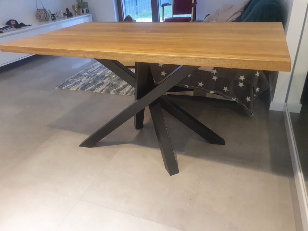 Stół z blatem z jasnego drewna i nowoczesną podstawą z czarnych, krzyżujących się metalowych elementów, na tle jasnej podłogi i fragmentu wnętrza mieszkania.