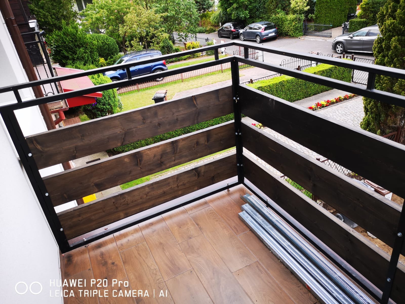 Barierka balkonowa z ciemnego drewna i czarnej stali, widok z góry na fragment balustrady z widocznymi elementami montażowymi i rurami leżącymi na podłodze balkonu, w tle zieleń i zabudowa...