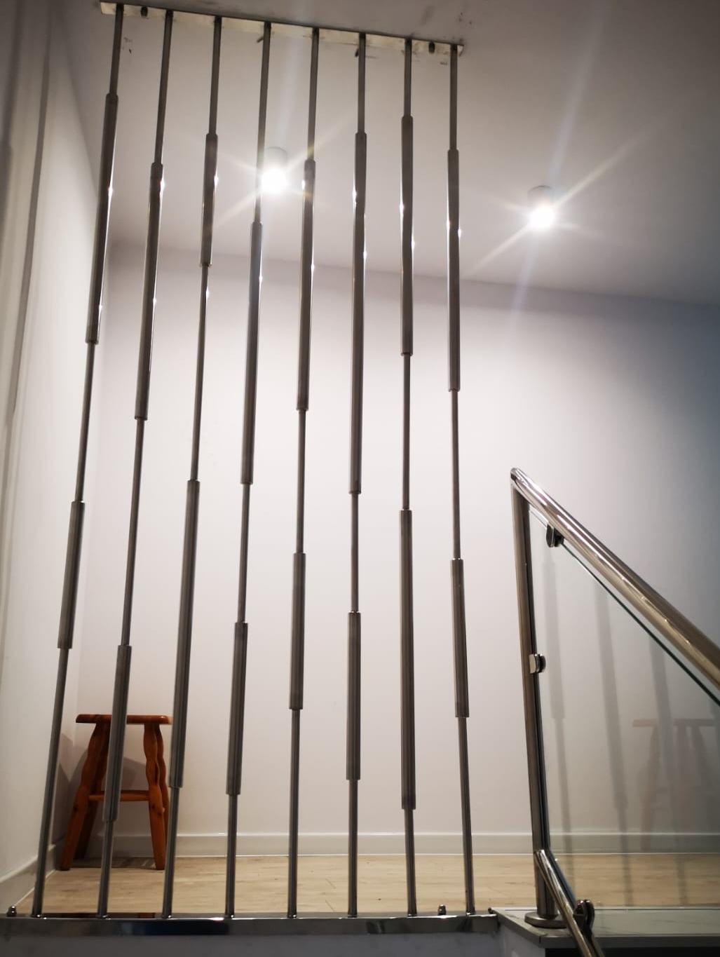 Metalowa balustrada schodowa z pionowymi prętami i szklanym panelem, widok z dołu na schody, z drewnianym stołkiem w tle.