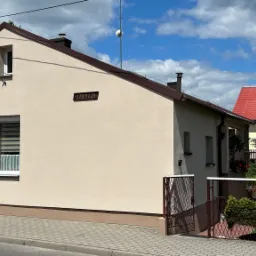 Jasnobeżowy dom jednorodzinny z brązowym dachem, widoczny z boku, z numerem 'Lesna 24' na ścianie, na tle błękitnego nieba z obłokami.