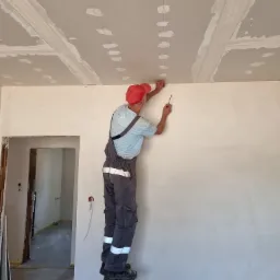Pracownik w czerwonej czapce na drabinie, wyznaczający linię na suficie z płyt kartonowo-gipsowych, widoczne szpachlowane łączenia płyt.