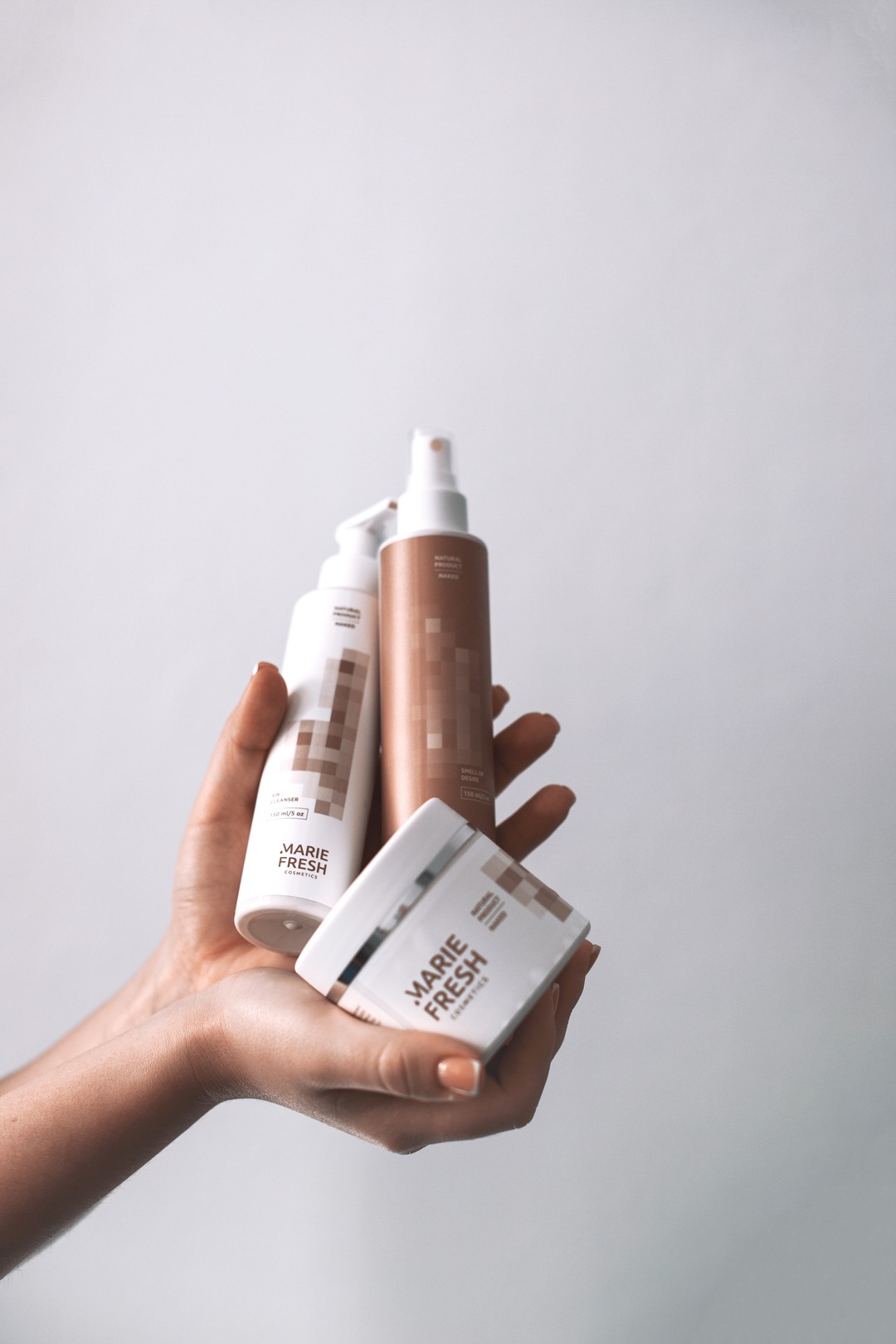 Dłoń trzymająca trzy produkty kosmetyczne marki Marie Fresh: krem w słoiczku, butelkę z pompką i spray, wszystkie w kolorystyce bieli i beżu z pikselowym wzorem.